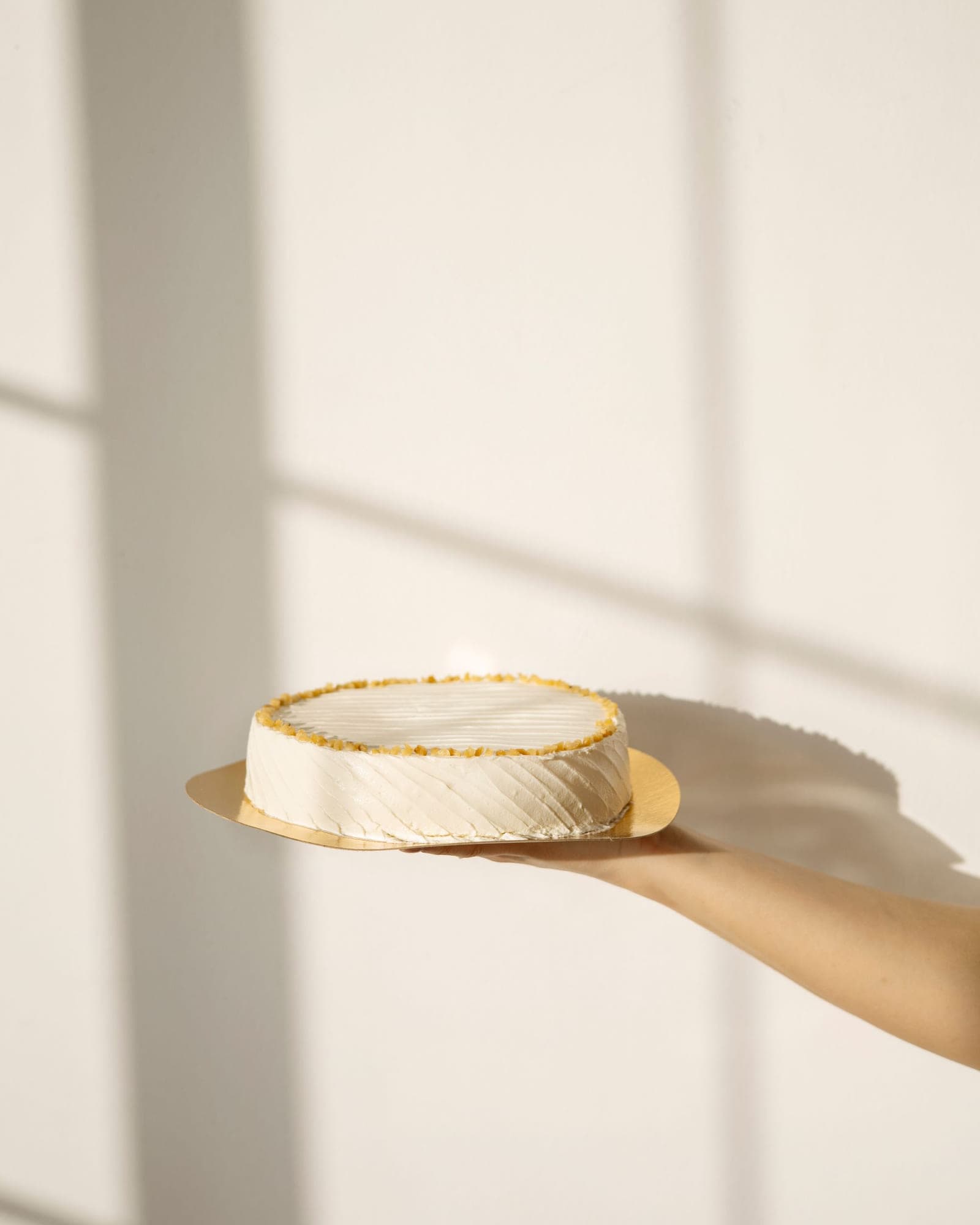 Hand holding a minimal ivory cake against a sunlit cream wall — presentation photo